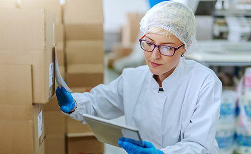 Caucasian blonde employee in sterile uniform crouching next to boxes and dealing with logistic of products. Food factory interior. In hand tablet.